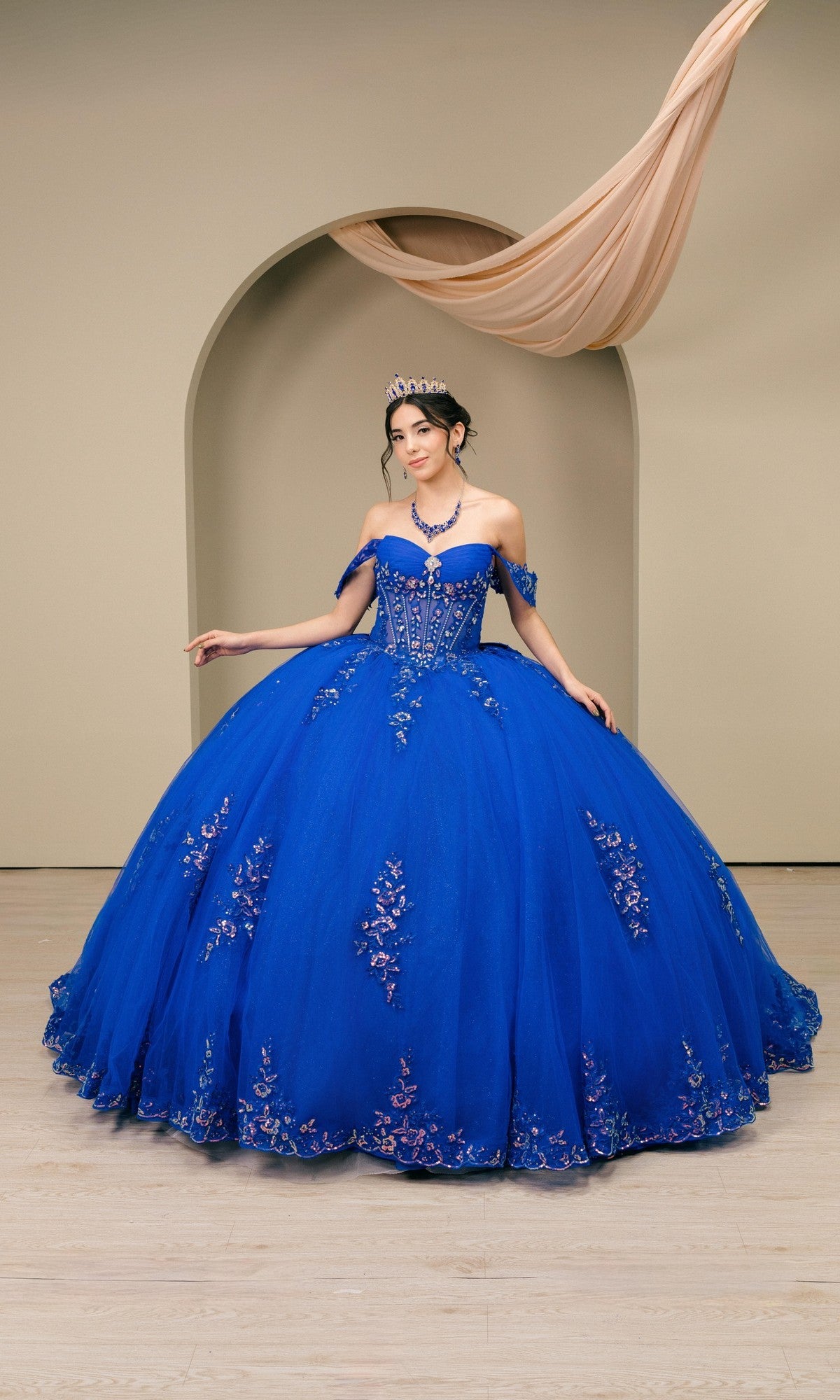 A woman in the Quinceanera Dress 8405JQ by Cinderella Couture in royal stands gracefully before an arched backdrop, wearing a sparkling tiara and showcasing floral applique details.